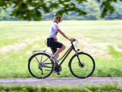 Kobieta w kasku jedzie na rowerze, który może być zarówno rowerem crossowym, jak i trekkingowym, po ścieżce w parku.