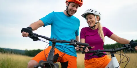 Uśmiechnięta para w kaskach i sportowych strojach, gotowa na przejażdżkę rowerami elektrycznymi. Wygodne akcesoria do roweru elektrycznego zapewniają komfort.