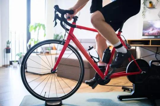 Intensywny trening na trenażerze kolarskim w domu. Czerwony rower, sportowiec w spodenkach i koszulce, butelki z wodą.