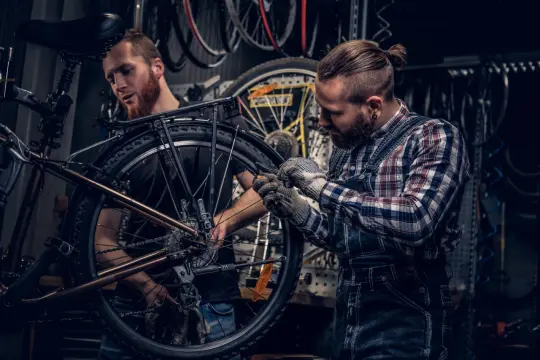 Dwóch mechaników pracuje nad rowerem w warsztacie. Jeden z nich przygląda się przerzutce, drugi dokręca śrubę. Profesjonalny serwis rowerowy.