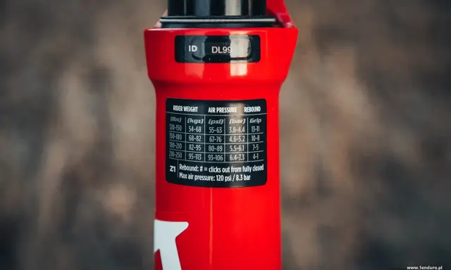 Czerwiony amortyzator Rock Shox z naklejką zawierającą tabelę ciśnień i ustawień powrotu dla różnych wag rowerzysty.