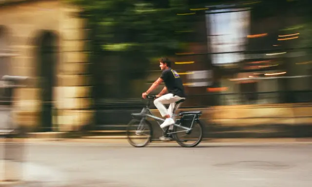 Mężczyzna na rowerze typu s-pedelec pędzi ulicą, rozmyte tło sugeruje dużą prędkość.