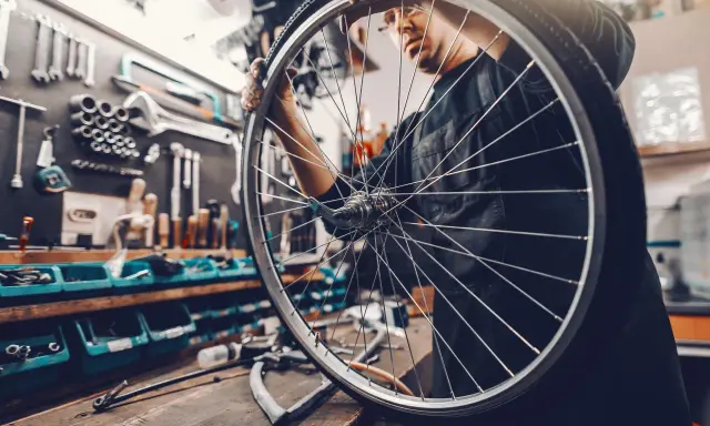 Mechanik sprawdza układy w rowerze, trzymając koło. W tle warsztat z narzędziami.