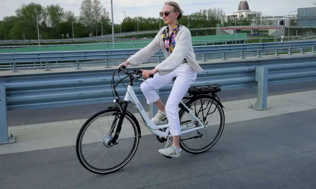 Młoda kobieta w białych spodniach i jasnym swetrze jedzie na rowerze elektrycznym, ciesząc się jazdą i osiągając swoją maksymalną prędkość.