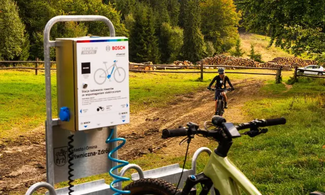 Stacje ładowania rowerów elektrycznych w Piwnicznej-Zdroju. W tle rowerzystka na szlaku.