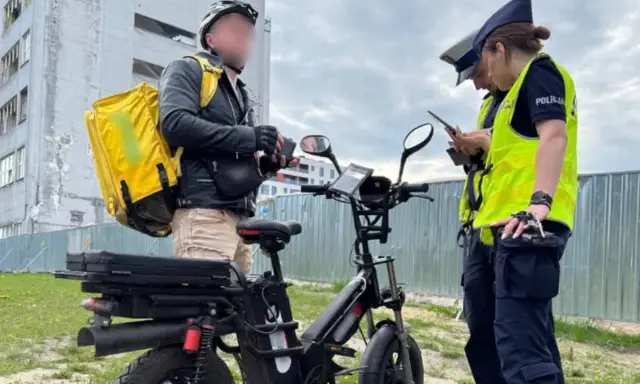 Dostawca z żółtą torbą na plecach rozmawia z policjantkami obok swojego roweru elektrycznego bez pedałowania.